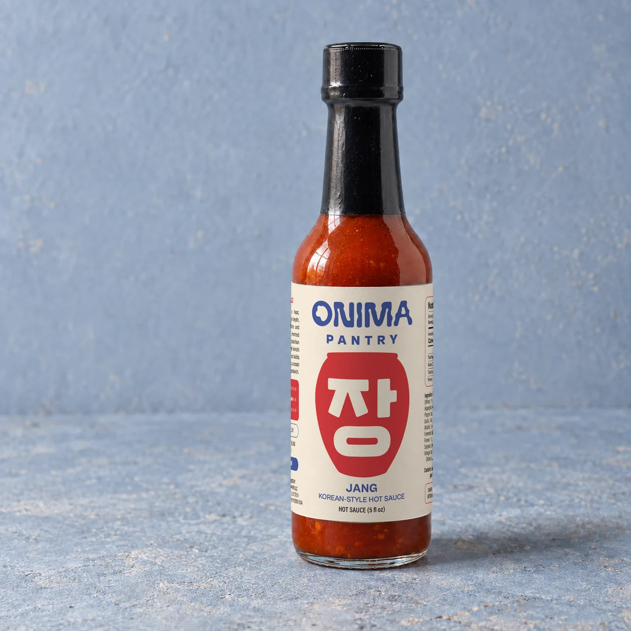 ONIMA JANG hot sauce bottle with its cream, blue and red label on a textured blue surface.