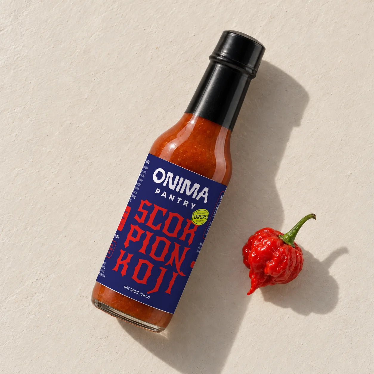 ONIMA Scorpion Koji hot sauce bottle with its navy and red label, lying beside a red pepper.