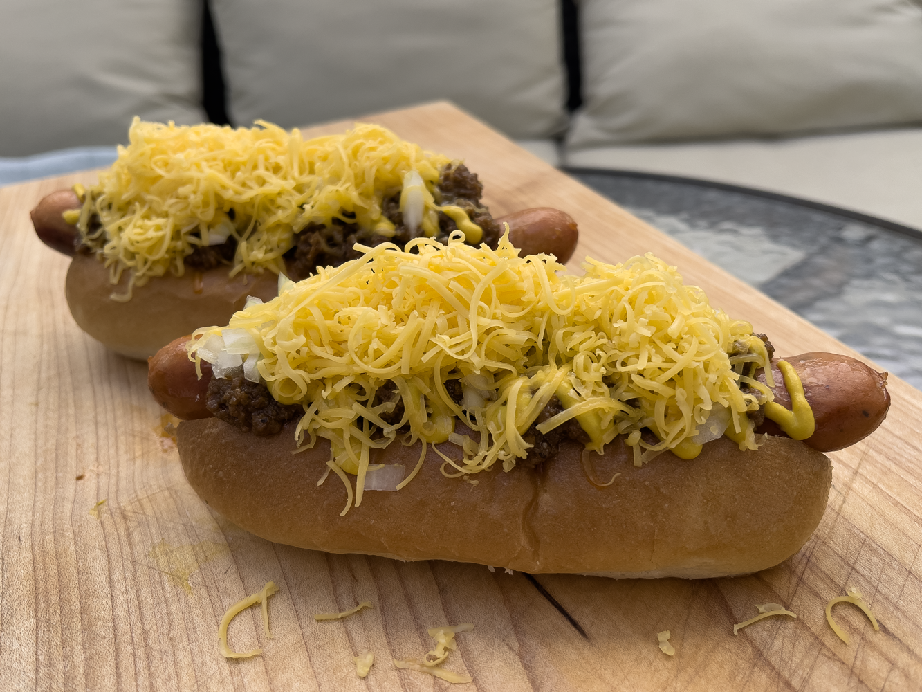 two chili dogs, Cincinnati style, topped with chili, onions, and cheese, sitting on a wooden cutting board
