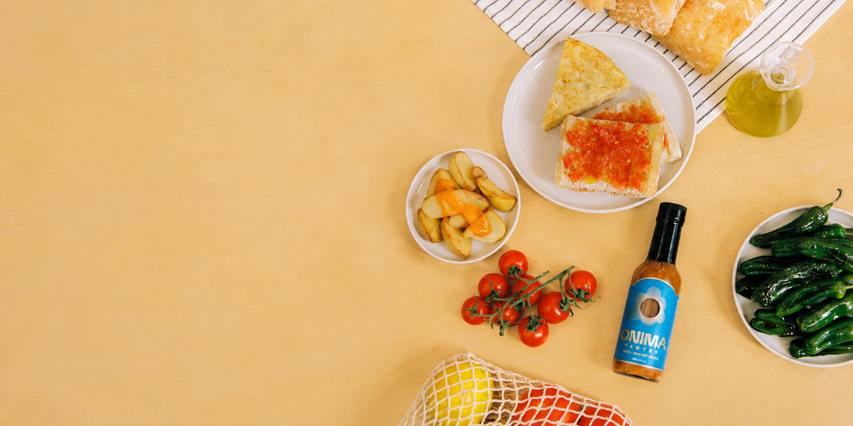 ONIMA hot sauce with tomatoes, padron peppers and tomato toast on a sunny yellow table