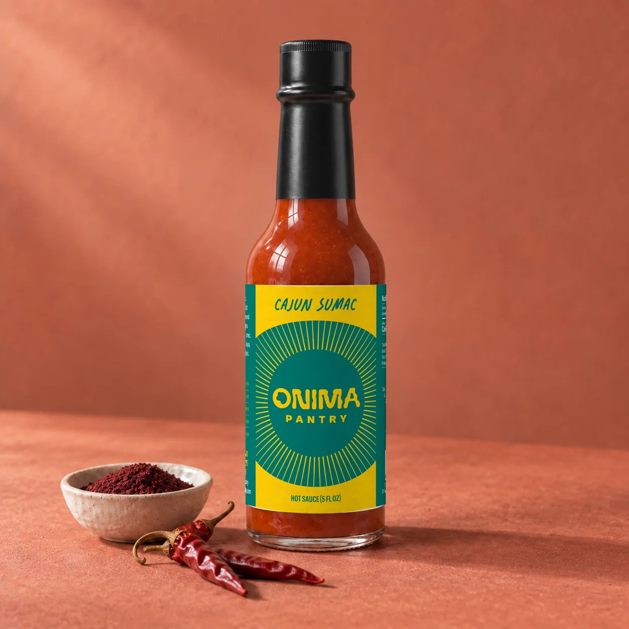 ONIMA Cajun Sumac hot sauce bottle with its teal and yellow label, beside sumac and dried peppers.